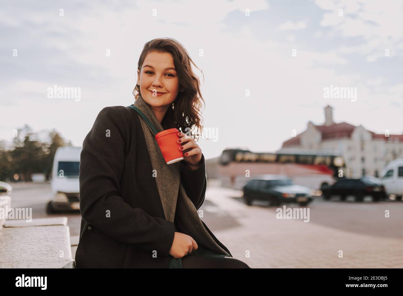 Vita su di sorridente bella femmina seduta su gradini mentre tenere una bevanda calda con l'auto sullo sfondo Foto Stock