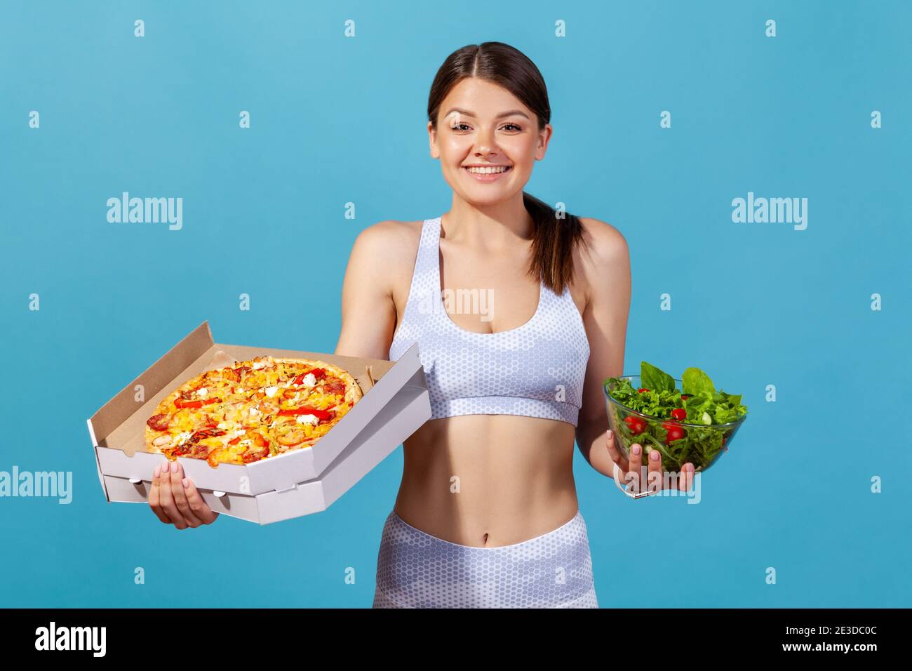 Donna atletica sorridente in sportswear bianco che tiene la scatola della pizza e ciotola con insalata fresca succosa, sognando circa il cibo della roba di rifiuto, mantenendo la dieta. Studio interno sh Foto Stock