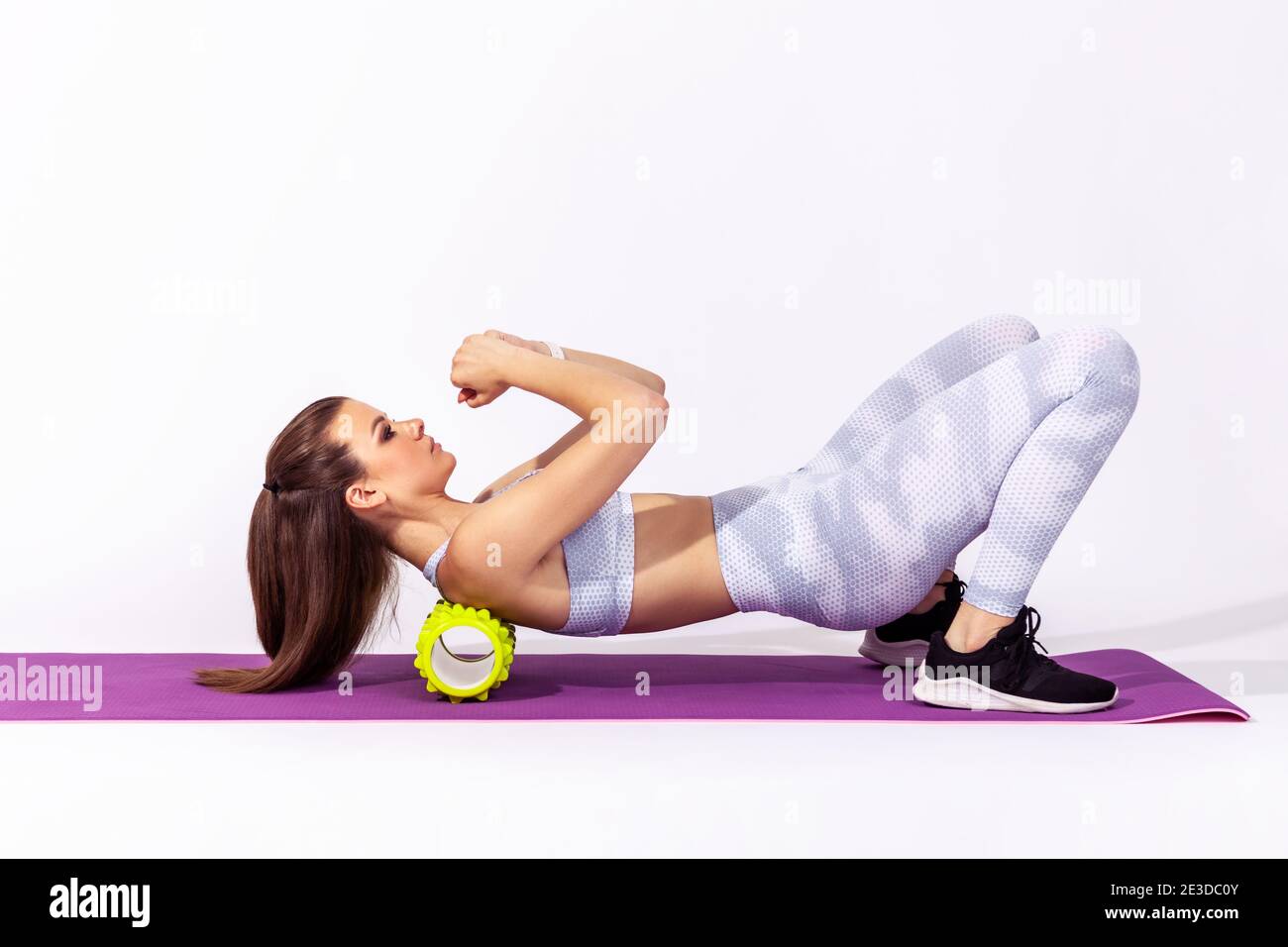 Vista laterale completa della donna atletica allenarsi sul tappetino yoga usando il massaggiatore a rullo in schiuma sotto le spalle, allungando i muscoli della colonna vertebrale, facendo esercizio con la fascia. Foto Stock
