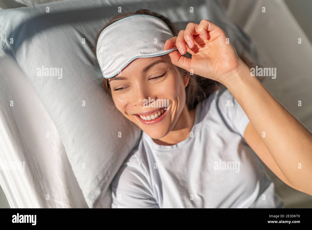 Donna asiatica felice svegliarsi sentendosi rinfrescato dalla maschera di sonno di bellezza occhio che dorme per una buona notte di riposo. Smiing giovane ragazza biraciale a letto. Sano Foto Stock