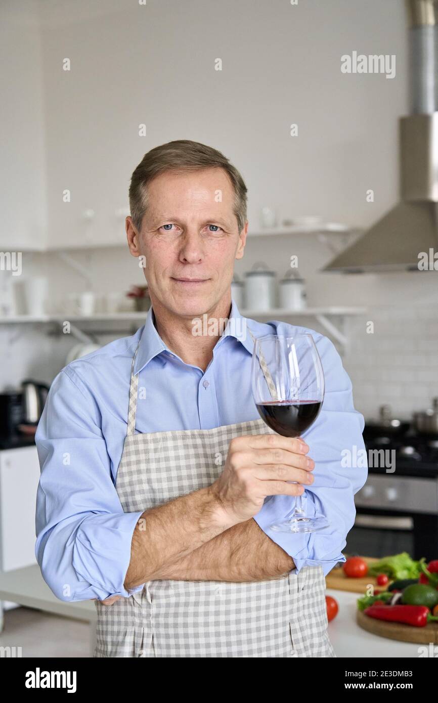 Uomo anziano sicuro di mezza età che tiene il bicchiere di vino in piedi in cucina, ritratto. Foto Stock