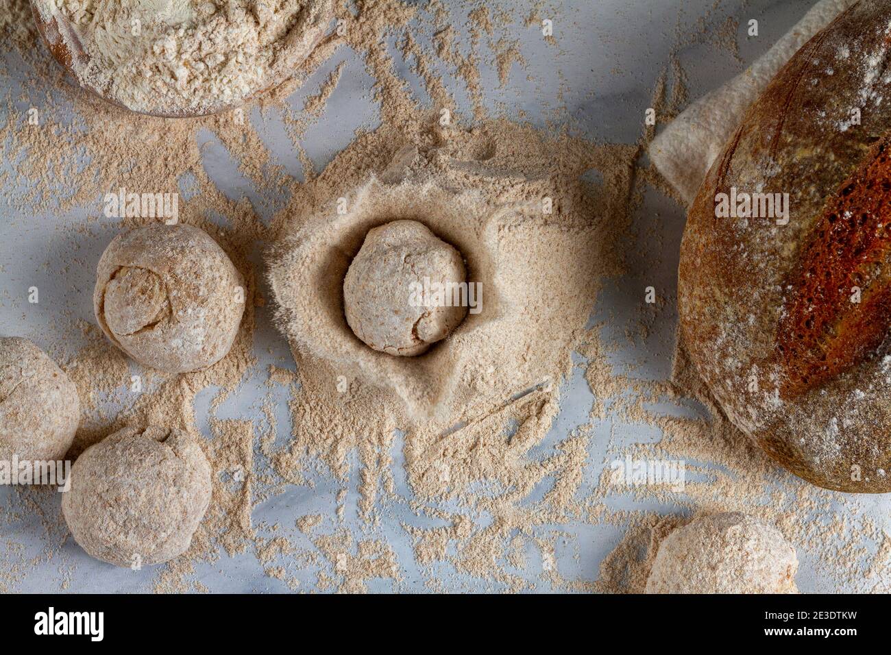 Immagine piatta di pani di pane, ingredienti di pasta, una ciotola di farina, un pane di pasta acida sul piano della cucina in marmo. Una focaccia di impasto viene fatta cadere su un pil Foto Stock