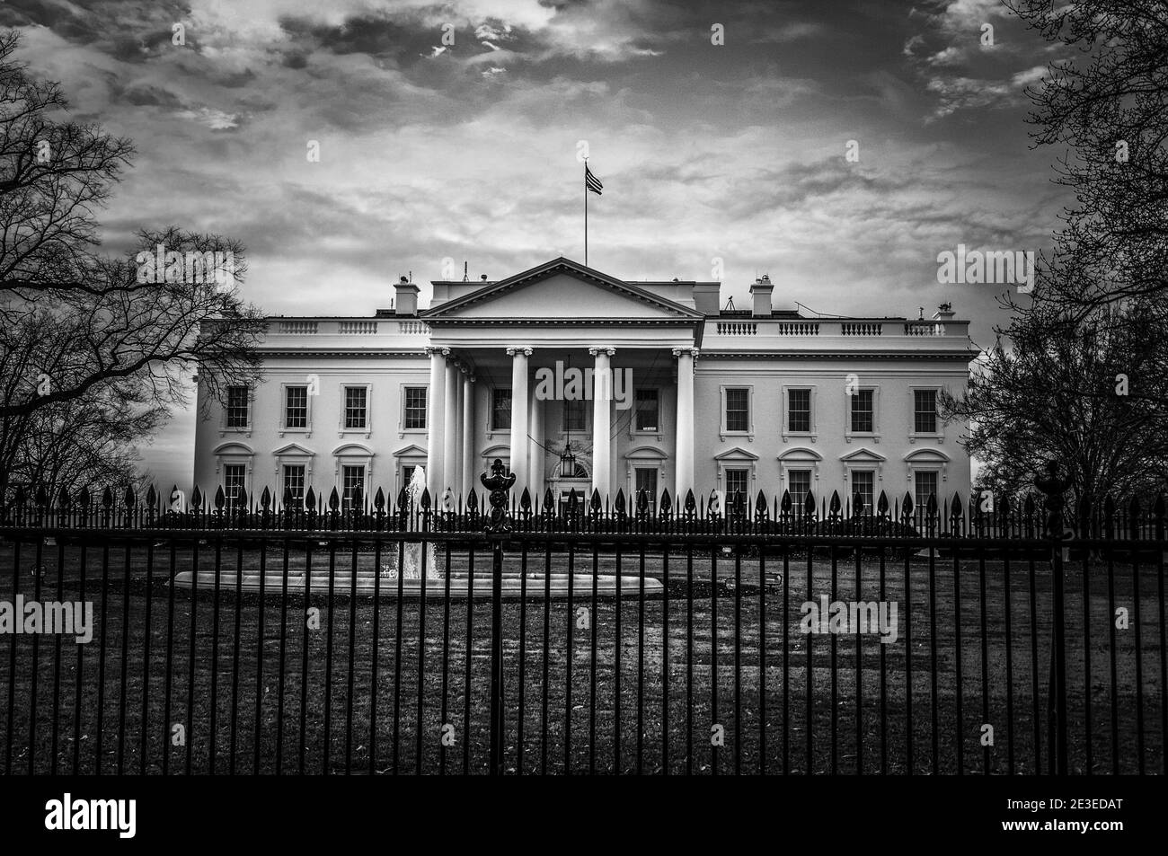 Vista in bianco e nero scuro della residenza presidenziale Whitehouse a Washington DC con cielo nuvoloso sullo sfondo e fontana, erba e recinzione in ferro Foto Stock
