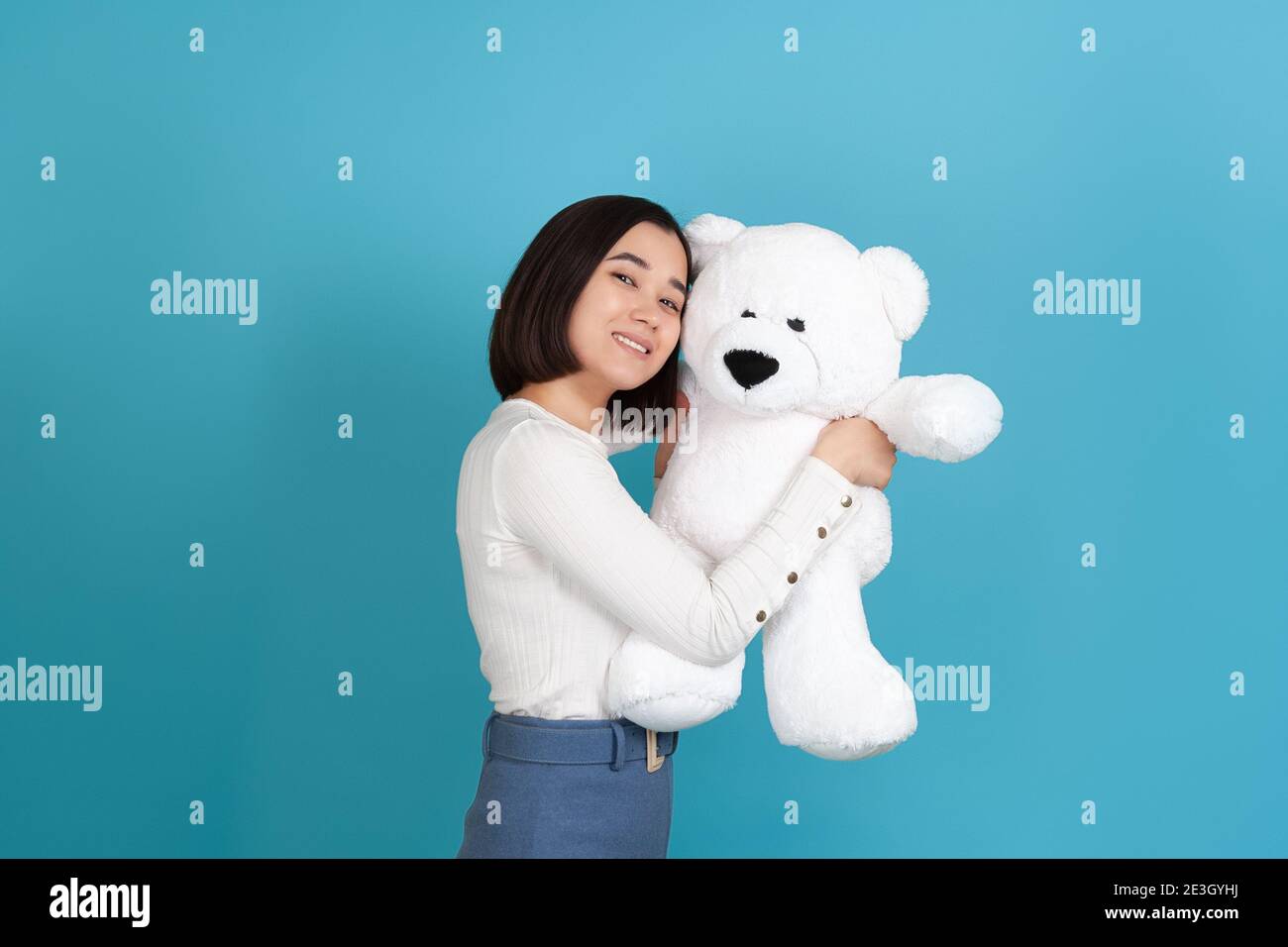 una donna asiatica molto gioiosa e gentile si alza lateralmente e si snuggisce fino ad un grande orsacchiotto bianco isolato su uno sfondo blu Foto Stock