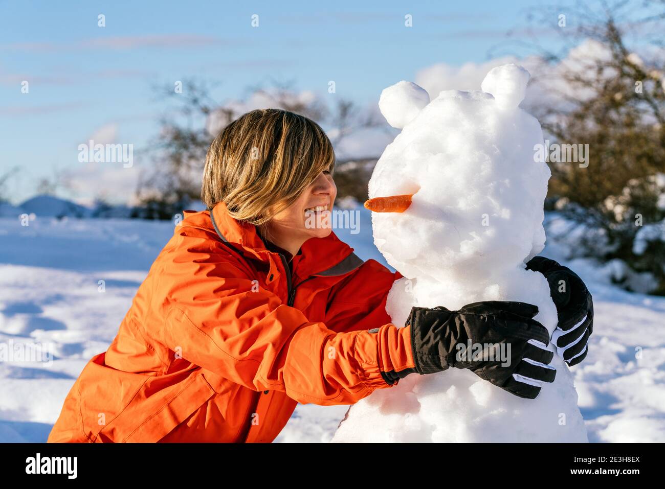 ritratto di una donna che tiene un pupazzo di neve con una carota sul naso su una montagna innevata con una bella paesaggio e indossare una giacca arancione facendo una v Foto Stock