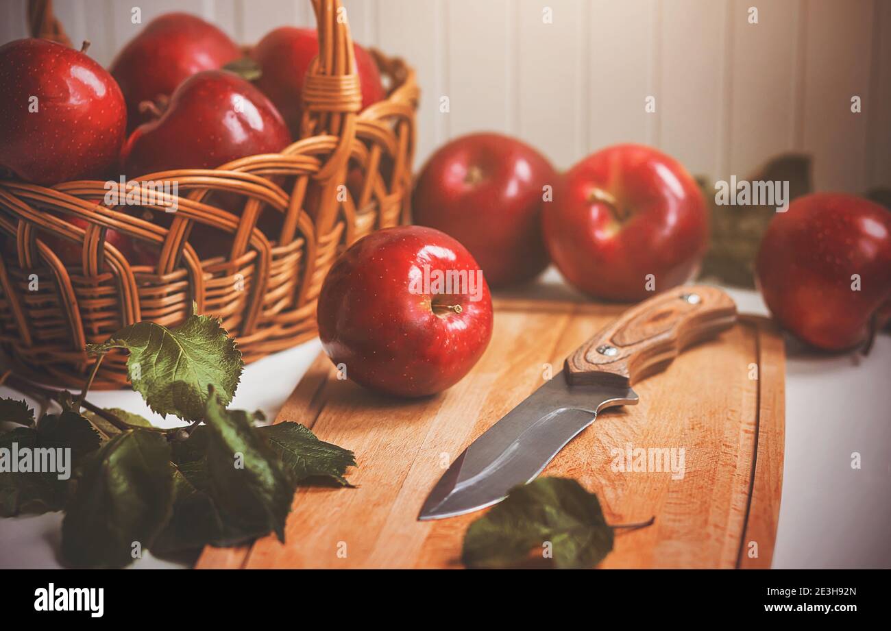 Le mele grandi rosse mature giacciono in un cesto di vimini e su un tagliere accanto a un coltello affilato. Uso della frutta raccolta. Vegetarianismo. Foto Stock