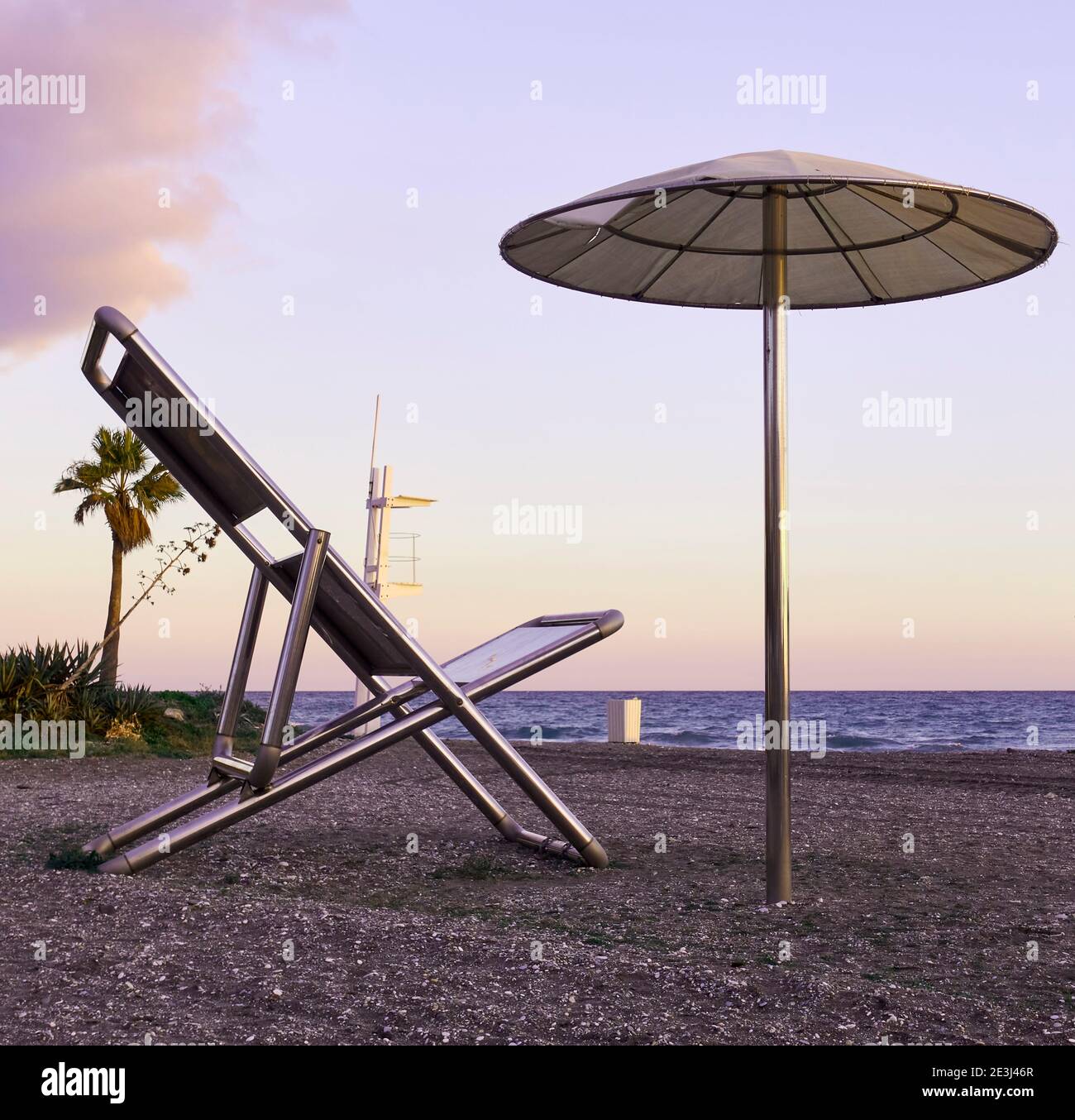 Colori del tramonto sul Mediterraneo da una gigantesca opera d'arte in acciaio di una sedia da spiaggia e un ombrello con un albero di palma, un'agave spike pendente e una torre di bagnino Foto Stock