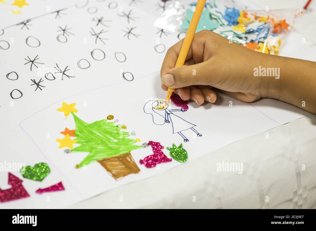 Concorso di disegno natalizio per bambini. Mano che tiene una penna di feltro gialla, foglio di carta con un disegno che raffigura una donna e un cane vicino ad una t di Natale Foto Stock