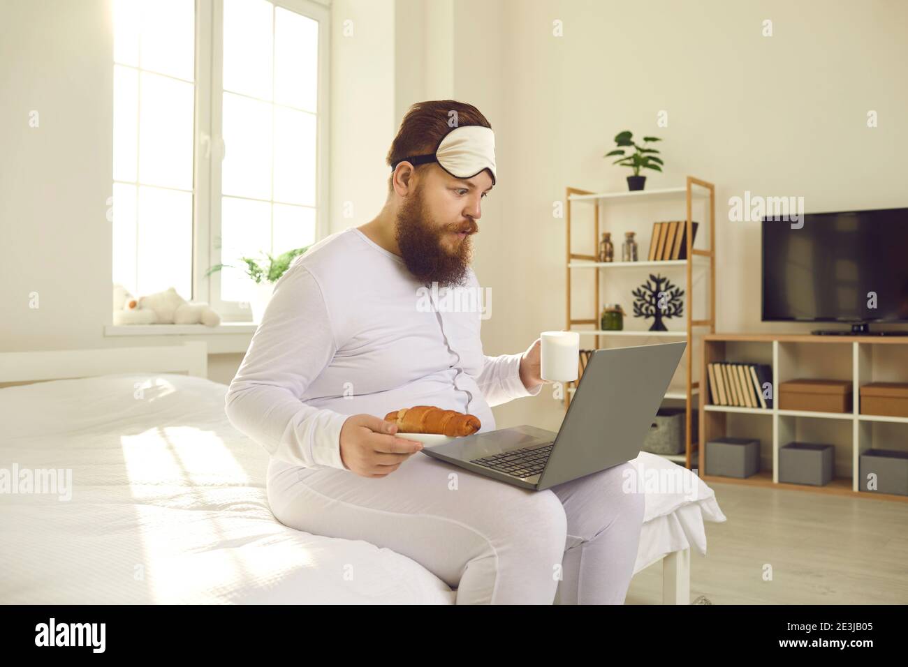 Uomo divertente in pigiami bianchi seduti con caffè e croissant a letto e guardando il computer portatile Foto Stock