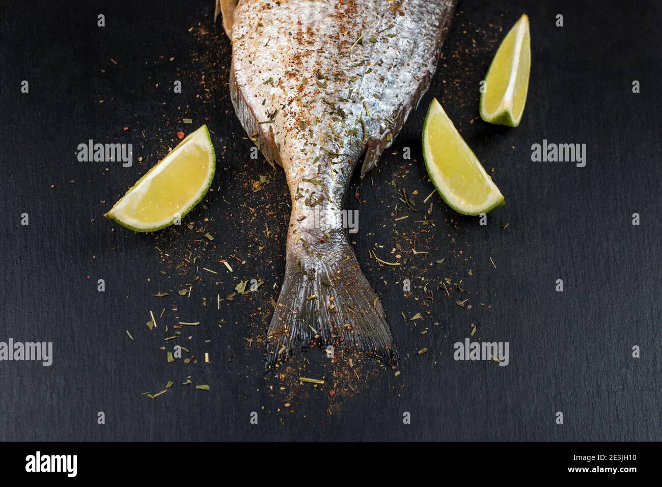 Coda di dorado fresco crudo su tavola di ardesia nera con spezie, erbe, limone e pepe rosso caldo. Pesce orata pronto per la cottura. Vista dall'alto Foto Stock