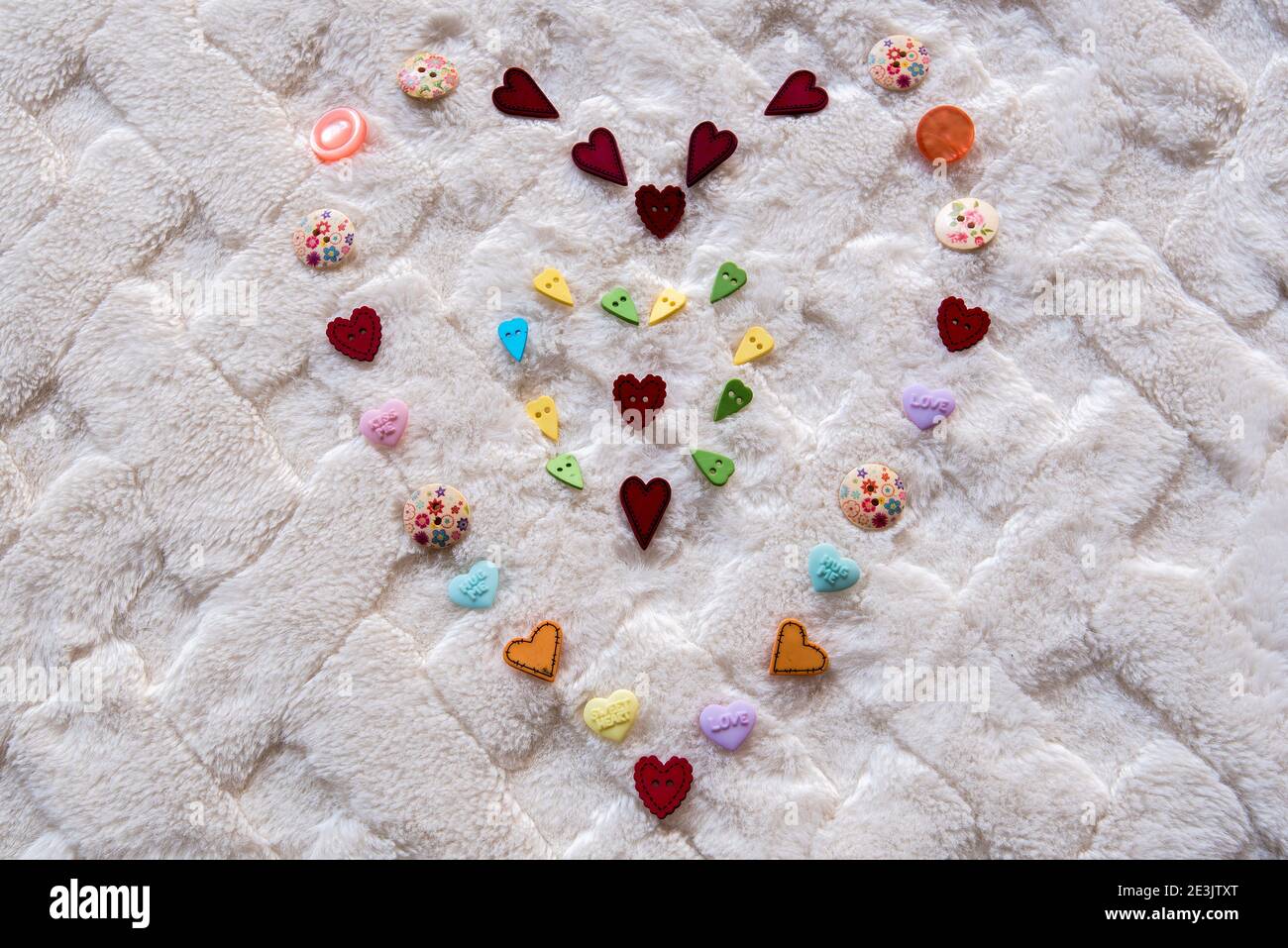 Design a cuore multicolore su una morbida coperta bianca. Il design è realizzato principalmente con bottoni a forma di cuore. Foto Stock