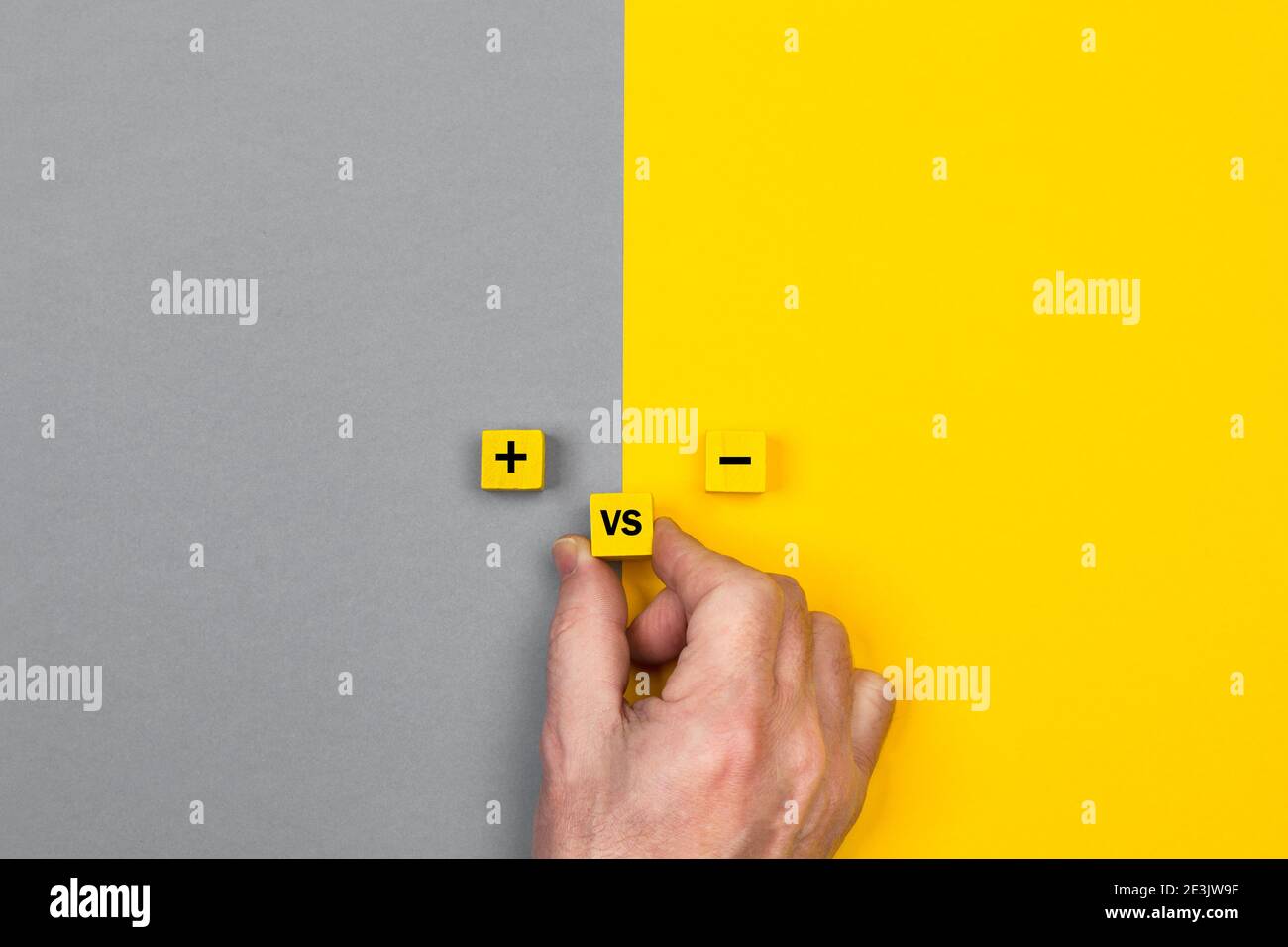 Uomo che tiene blocchi di legno con segno meno e più e versus su uno sfondo giallo e grigio Foto Stock