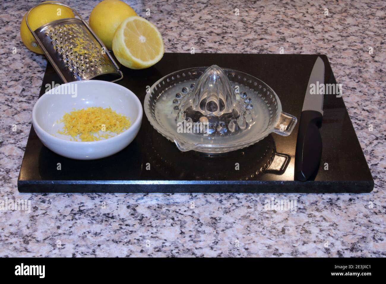 Spremiagrumi in vetro e grattugia di scorza di limone con limoni e scorza  di limone in un piccolo recipiente su un pannello di granito nero. Cottura  ordinata e preparazione del cibo Foto