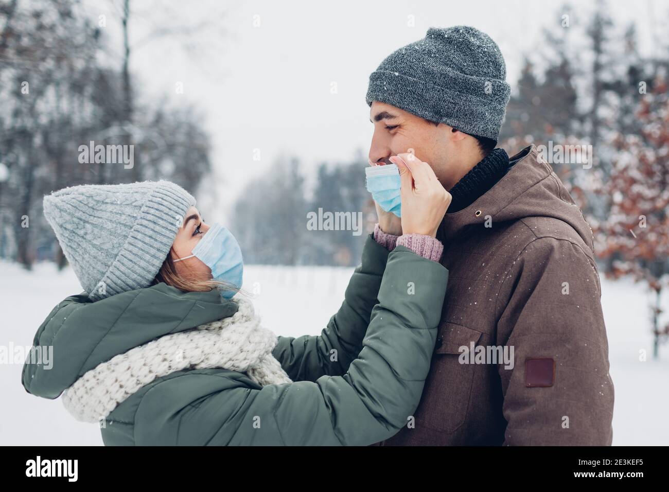 Donna mette la maschera sul viso del ragazzo in un parco invernale innevato. Coppia in amore camminare all'aperto. Misure di sicurezza per la pandemia di coronavirus Foto Stock
