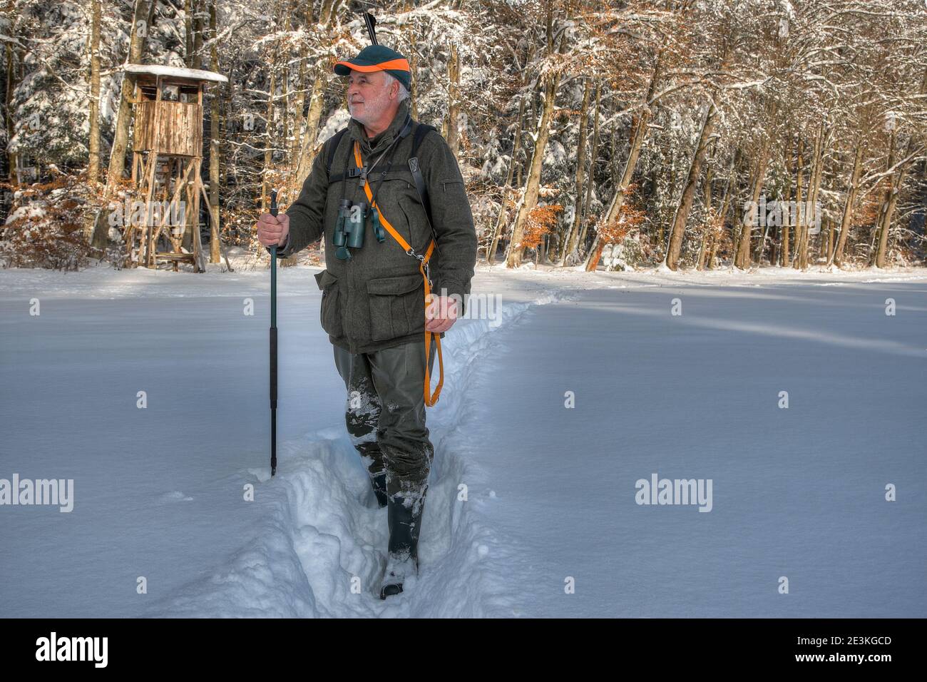 In un bel pomeriggio invernale, un cacciatore sta stalking nella sua zona di caccia e cammina attraverso la neve fresca e profonda. Il suo pulpito di caccia si trova dietro di lui dentro Foto Stock