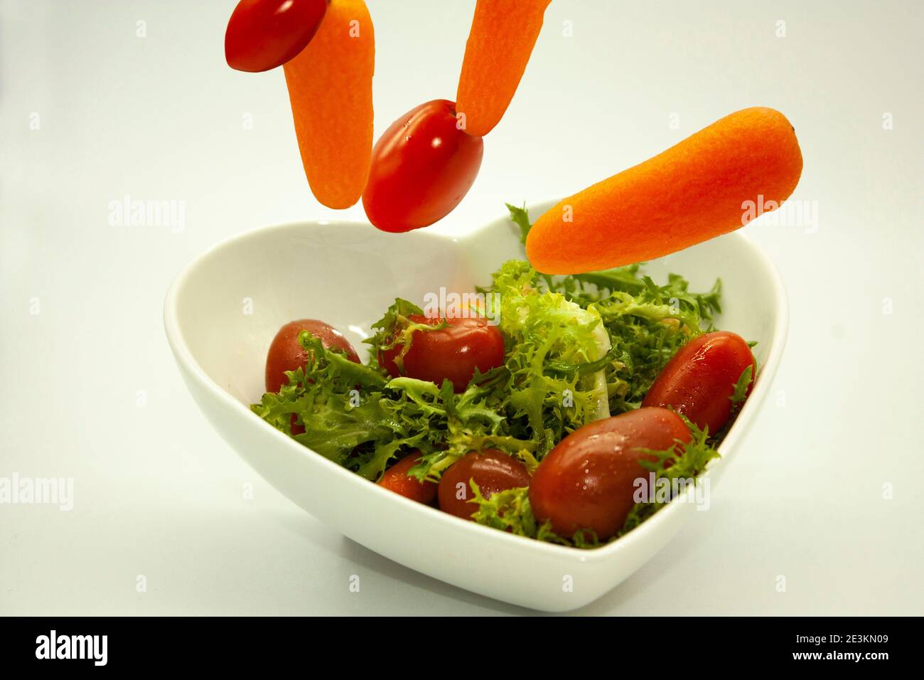 Verdure che cadono in ciotola a forma di cuore con insalata vegana organica grezza combinazione di lecguga, pomodori ciliegia rossa, e carote Foto Stock