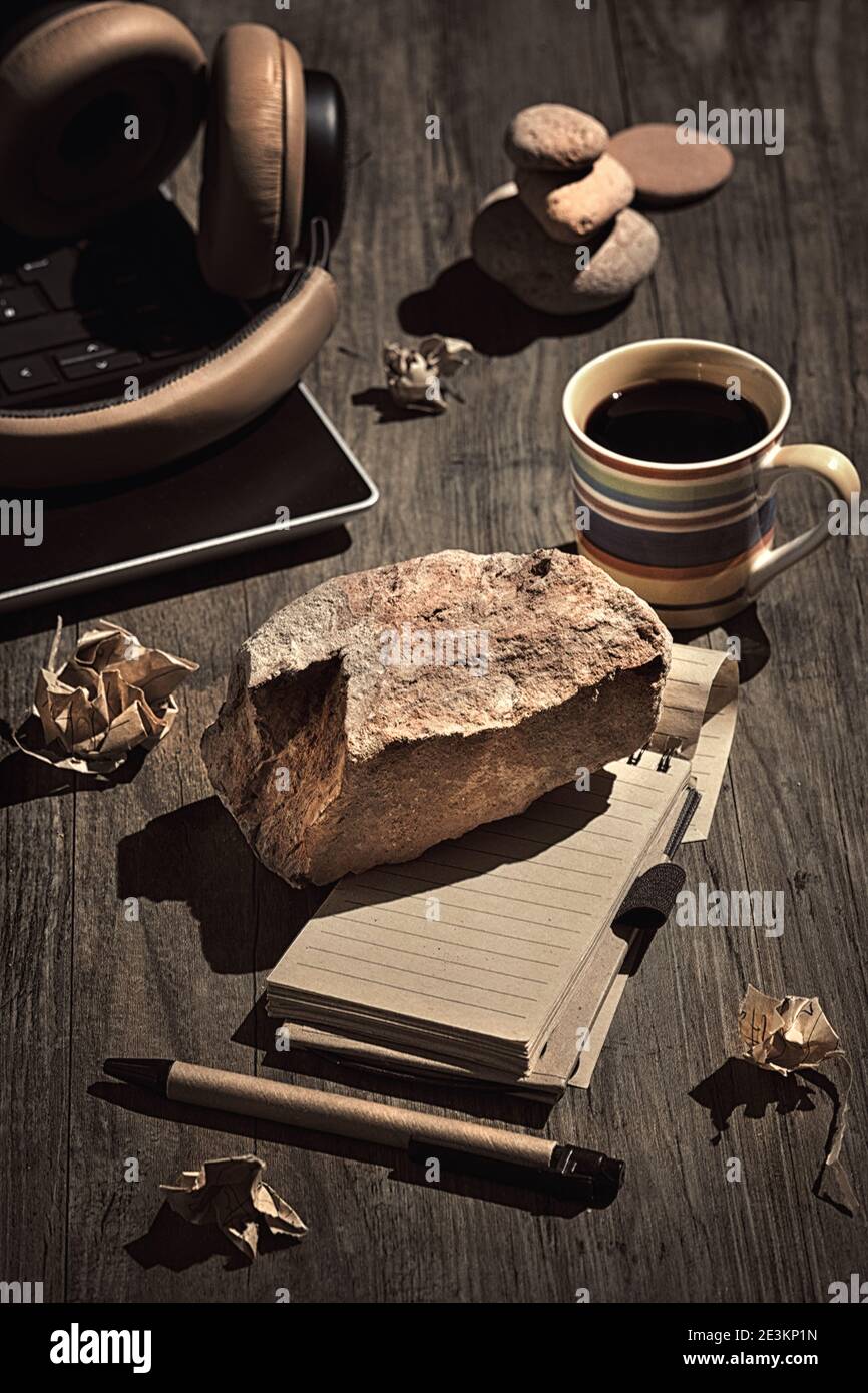 Blocco di scrittura. Concetto di vita morta, disposizione su tavolo di legno scuro. Pietra pesante, mattone su bianco aperto nota blocco. Computer portatile, auricolari, occhiali, pietra Foto Stock