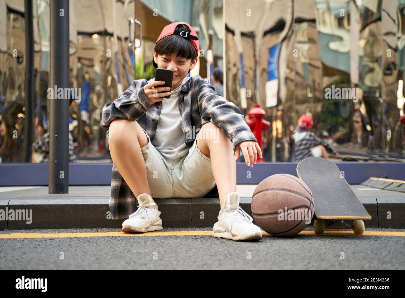 ragazzo asiatico adolescente con basket e skateboard seduto sul marciapiede di strada guardando il cellulare Foto Stock