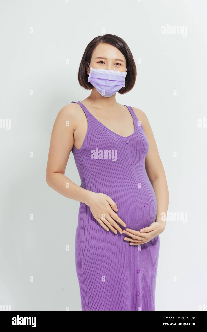 Donna incinta asiatica indossare maschera viso tenendo il ventre, guardare la fotocamera, isolato su sfondo bianco, covid-19 concetto Foto Stock