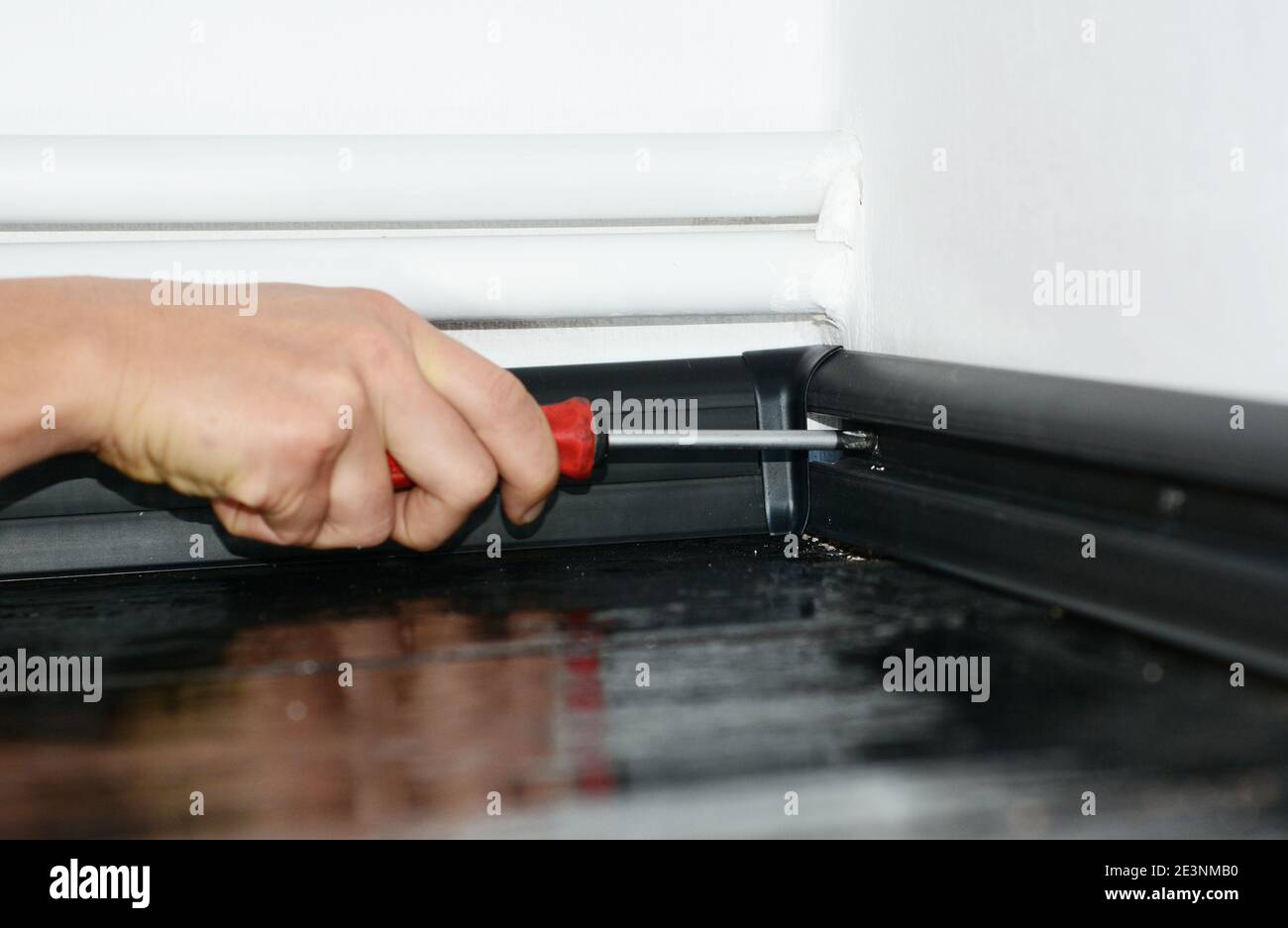 Un uomo sta installando un pannello di protezione in plastica nera su una parete bianca tappezzata nell'angolo di una stanza con tubi di riscaldamento centrali usando un cacciavite. Foto Stock