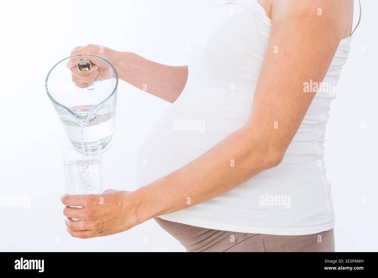 Donna incinta che versa acqua dal vaso al bicchiere su sfondo bianco Foto Stock