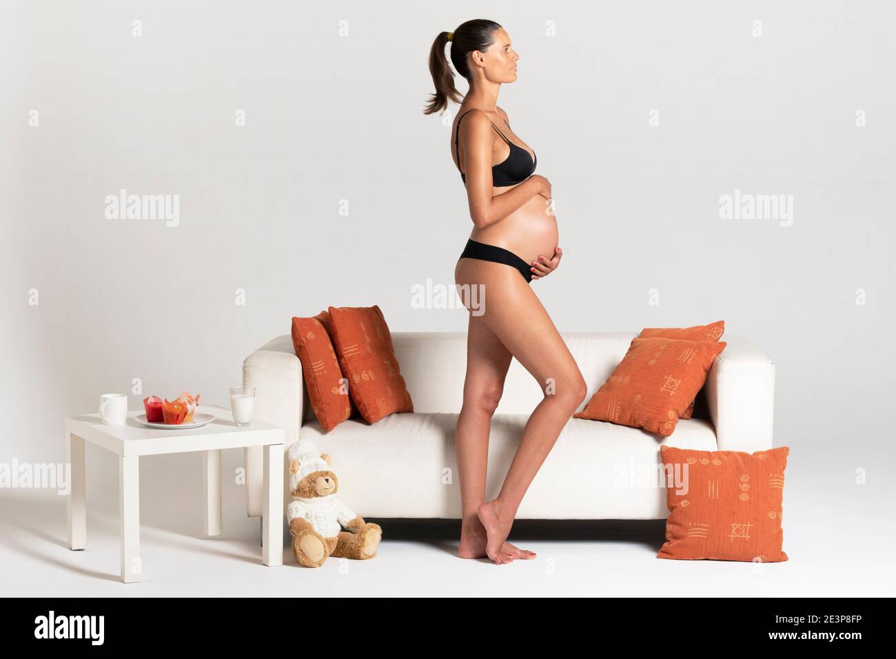 Giovane bella donna incinta in piedi vicino ad un divano bianco tenendo le mani sulla sua biancheria intima di vestirsi del ventre Foto Stock