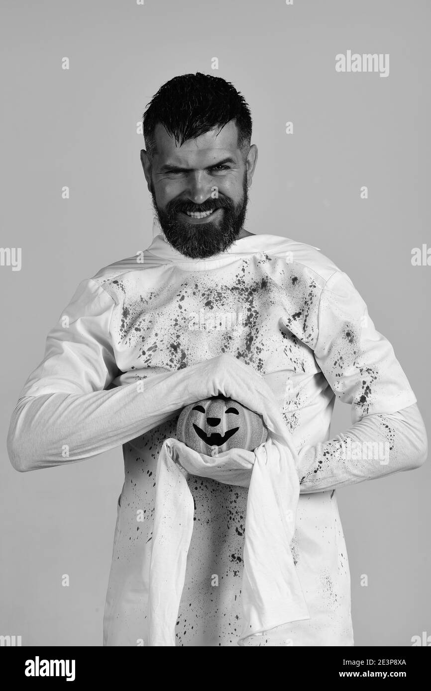 Halloween e concetto di vacanza felice. Personaggio di Halloween in costume fantasma bianco a maniche lunghe. Il ragazzo con la barba tiene la zucca arancione con il sorriso. Uomo con il sorriso malvagio sul viso isolato su sfondo grigio. Foto Stock