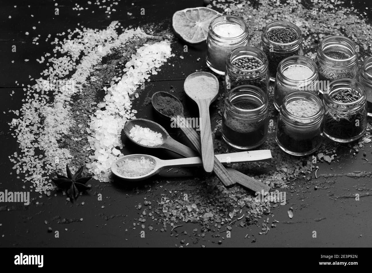 Cucchiai di legno con paprika, curcuma e sale marino vicino agli agrumi e spezie mescolano sul tavolo. Set di spezie su sfondo grigio scuro. Composizione di sale colorato e spezie vicino a vasi con erbe. Concetto di arte alimentare. Foto Stock