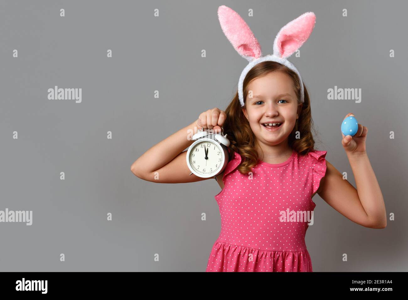 Carino ragazza felice in un costume di coniglio su uno sfondo grigio. Il  bambino tiene in mano una sveglia e un uovo di Pasqua Foto stock - Alamy