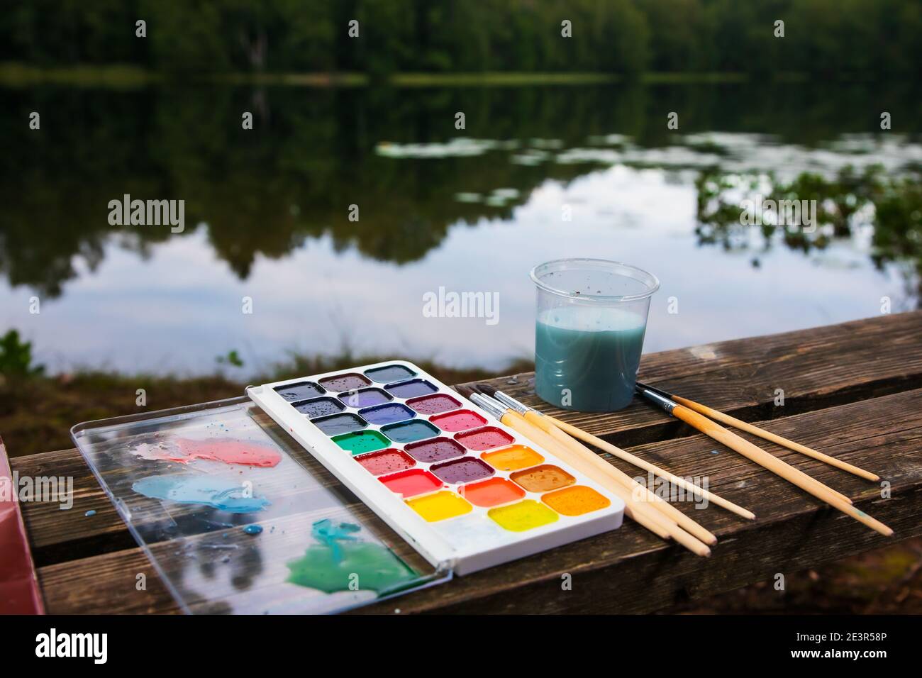 Primo piano della configurazione dell'acquerello con i pennelli e l'acqua della tavolozza di acquarelle vasetto adagiato su una panchina di legno vicino ad un lago forestale - concetto di scansione ad aria plein Foto Stock
