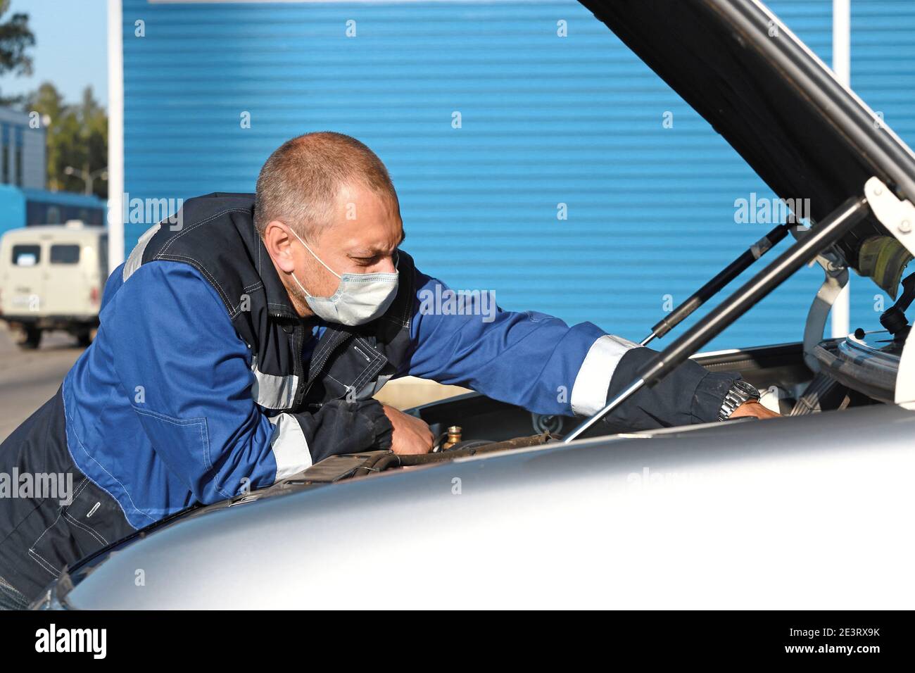 Un uomo bianco che indossa una maschera medica sul viso ripara un'auto all'esterno in uno spazio aperto. Foto Stock