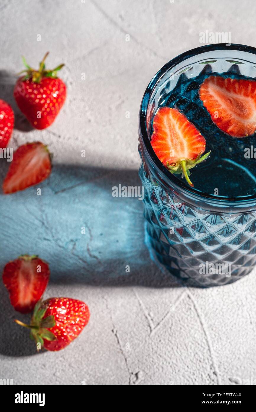 Una tazza di vetro blu geometrico con acqua fresca e frutti di fragola con colorati raggi di luce ombra su sfondo in pietra e cemento, vista angolare Foto Stock