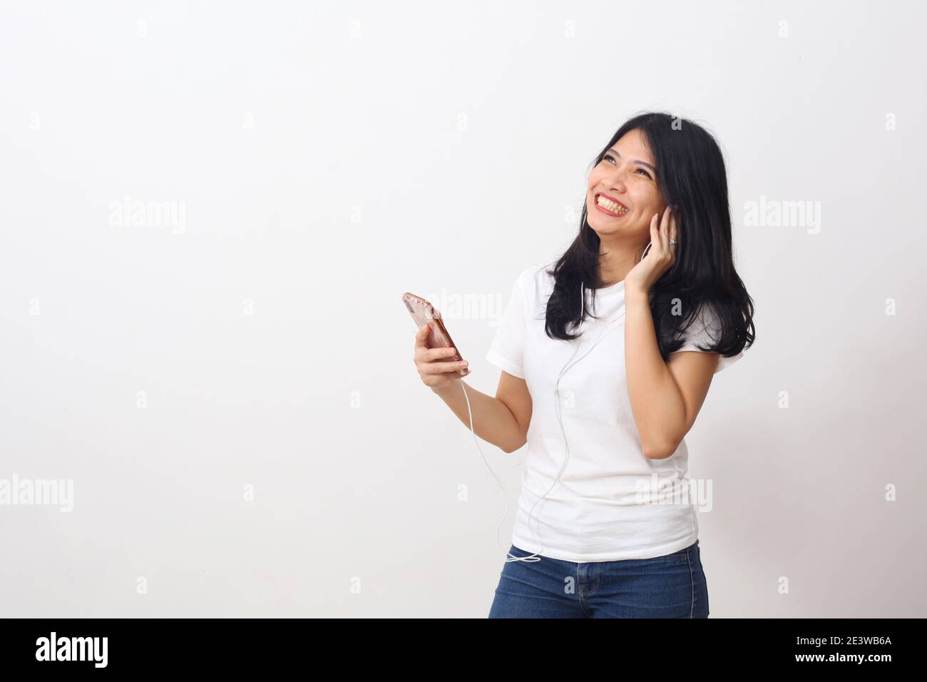 Bella giovane donna asiatica godere di ascoltare la musica o al telefono con le cuffie mentre in piedi e ridere. Isolato su bianco con copyspace Foto Stock