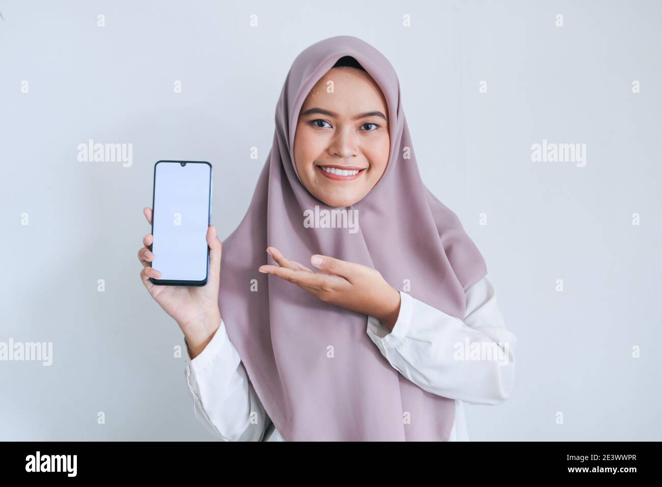 Giovane islam asiatico donna che indossa il copricapo sta puntando il dito sullo schermo bianco del telefono con sorriso e sensazione felice. Donna indonesiana sullo sfondo grigio Foto Stock