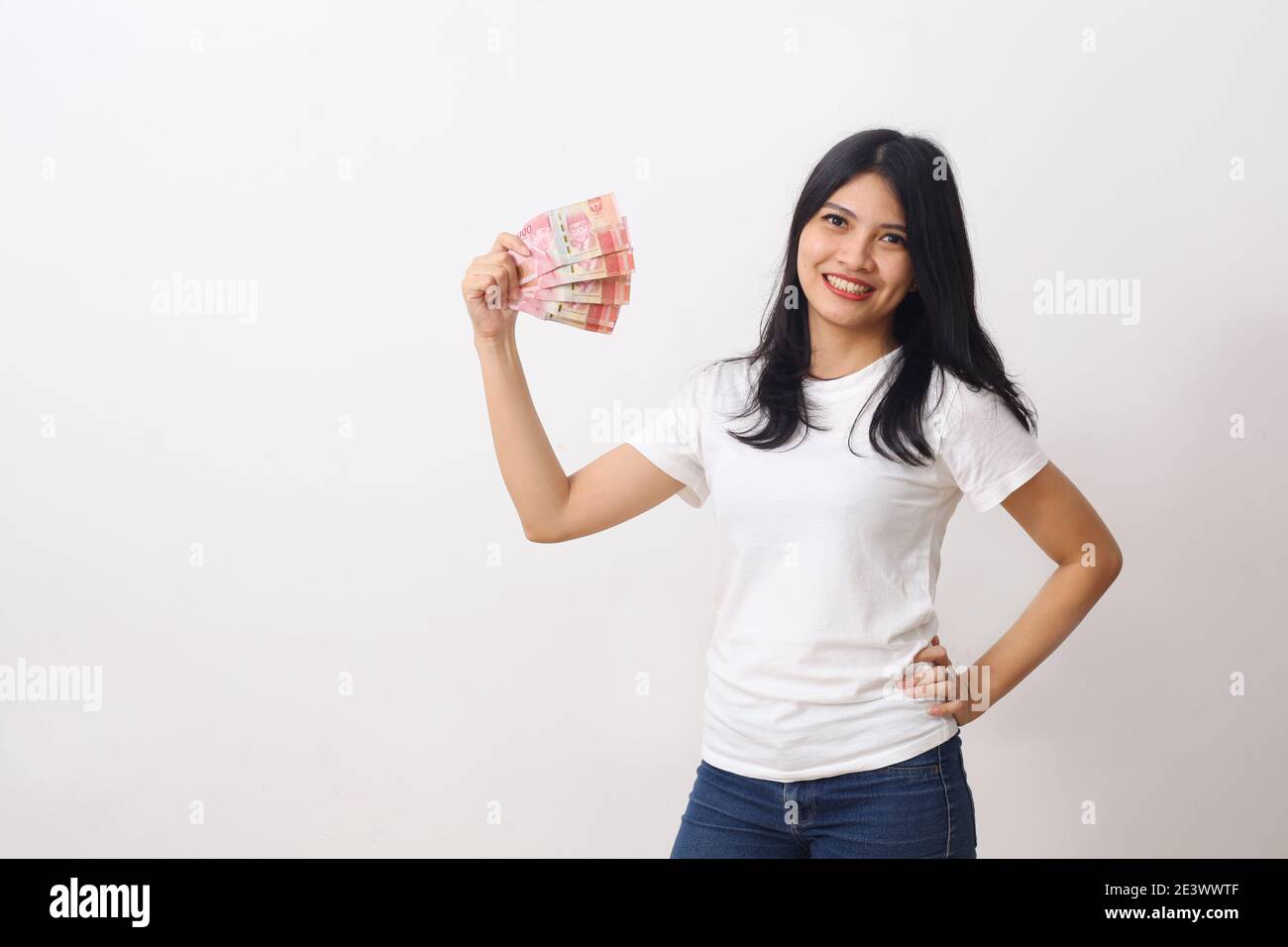 La ragazza asiatica felice tiene un denaro indonesiano mentre si è in piedi contro su parete bianca con copyspace Foto Stock