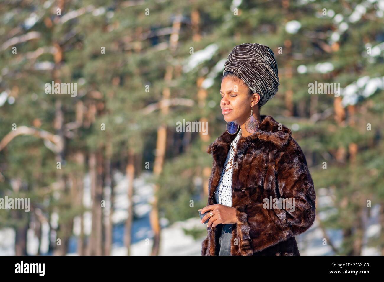 Vilnius, Lituania - Gennaio 20 2021: Bella ragazza con cappotto di pelliccia bere caffè caldo o tè sulla neve su un lago ghiacciato in inverno con alberi di foresta Foto Stock