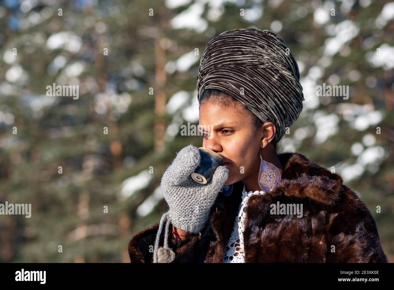 Vilnius, Lituania - Gennaio 20 2021: Bella ragazza con cappotto di pelliccia bere caffè caldo o tè sulla neve su un lago ghiacciato in inverno con alberi di foresta Foto Stock