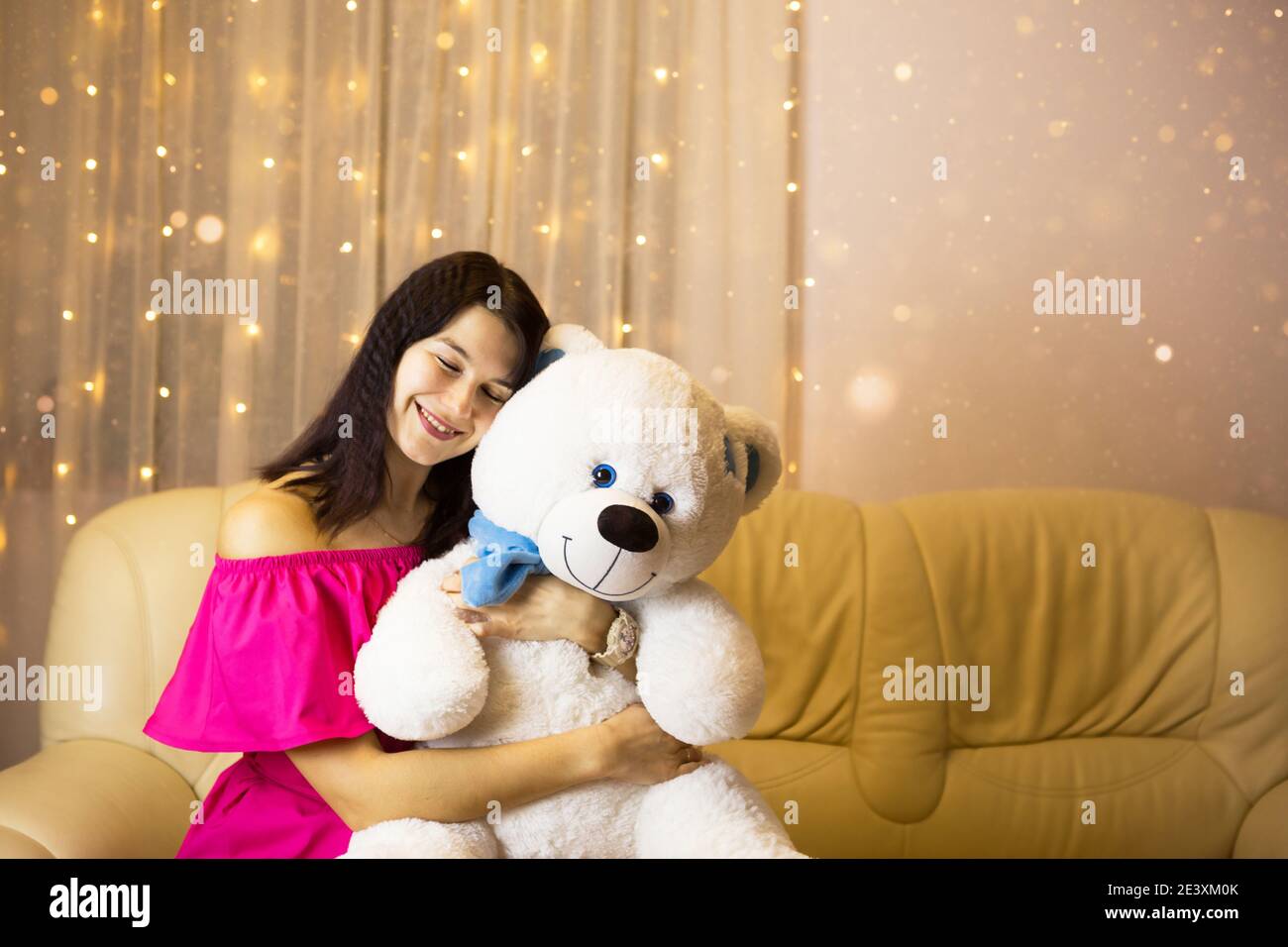 Ragazza felice in un vestito rosa abbracciando un grande orso bianco morbido a casa sul divano. Regalo da una persona cara, giorno di San Valentino, compleanno, vacanza. Spazio di copia Foto Stock