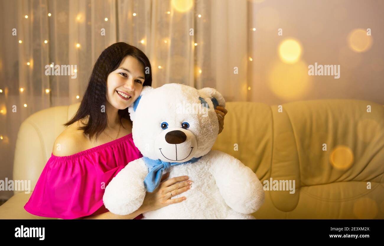 Ragazza felice in un vestito rosa abbracciando un grande orso bianco morbido a casa sul divano. Regalo da una persona cara, giorno di San Valentino, compleanno, vacanza. Spazio di copia Foto Stock