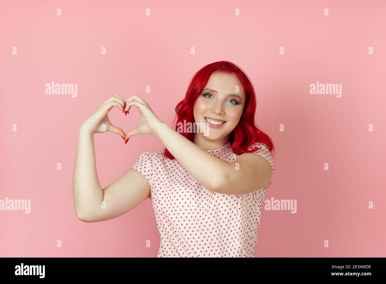 Primo piano una donna felice e ridente con i capelli rossi fa un segno del cuore dalle sue dita al suo fianco, isolato su uno sfondo rosa. Foto Stock