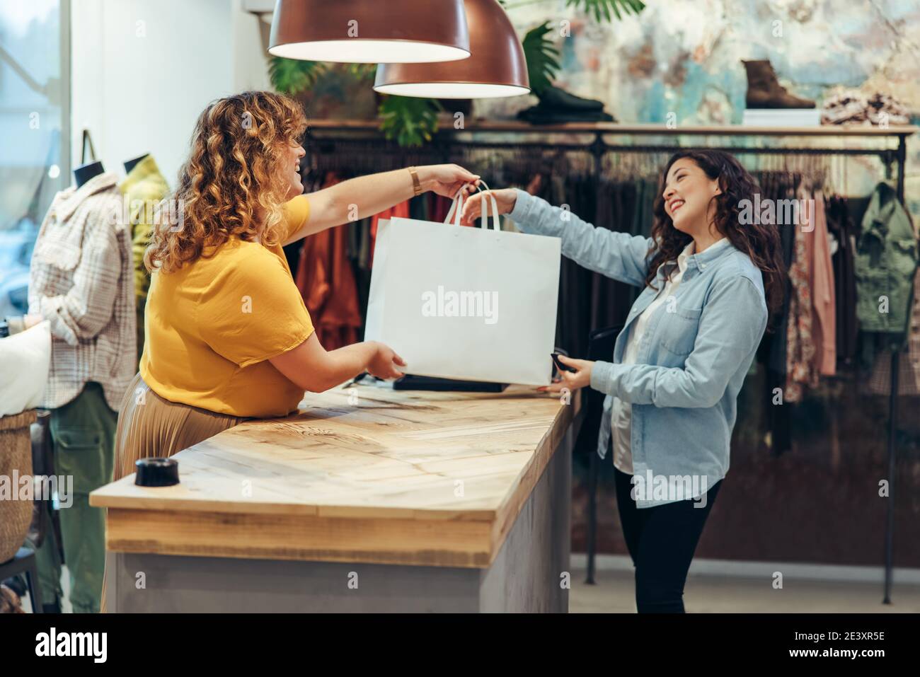 Venditore amichevole di negozio di abbigliamento che dà borse per la spesa a cliente femminile soddisfatto. Il proprietario del deposito di moda consegna la borsa di shopping ad un cus femminile Foto Stock