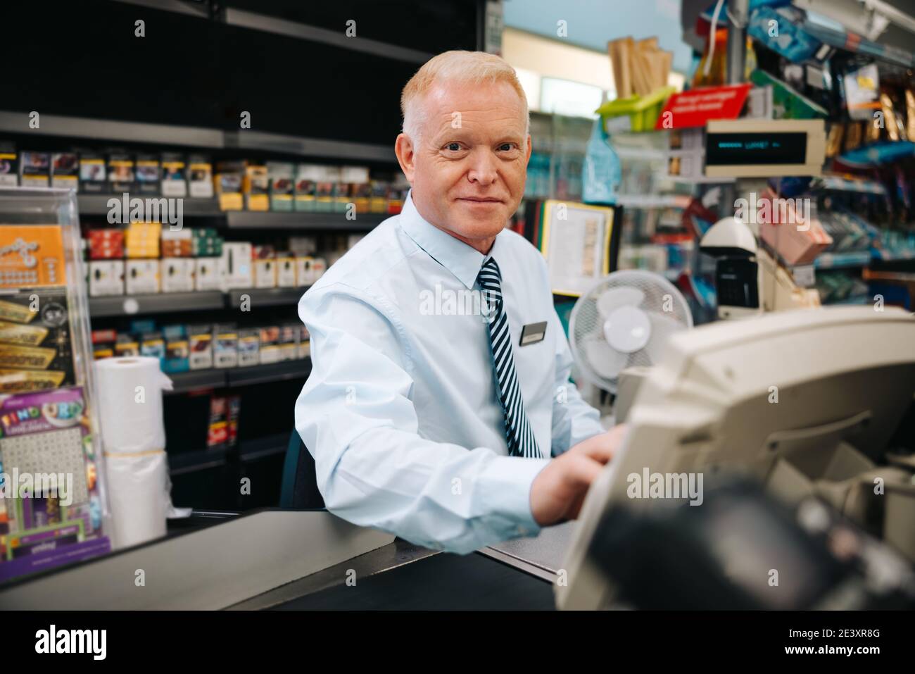 Uomo anziano seduto al super mercato checkout e guardando la fotocamera. Responsabile del negozio di alimentari al banco del check-out. Foto Stock