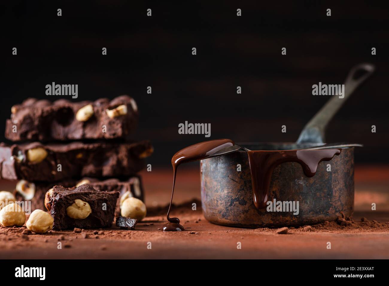 In primo piano alcuni pezzi accatastati di cioccolato fondente con nocciole. Oltre alla pentola di rame con il cioccolato fuso e il cucchiaio gocciolante Foto Stock