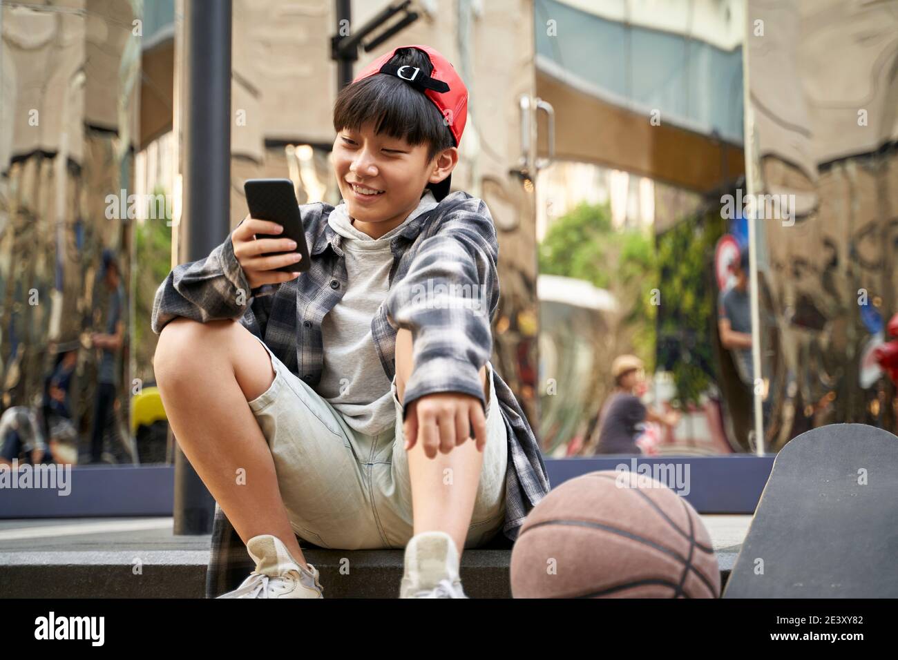 ragazzo asiatico adolescente con basket e skateboard seduto sul marciapiede di strada guardando il cellulare Foto Stock