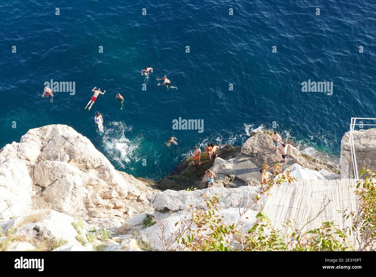 Croazia: Dubrovnik. Gruppo di giovani che nuotano nelle acque turchesi ai piedi del bar Buza e dei bastioni orientali della città vecchia, Foto Stock