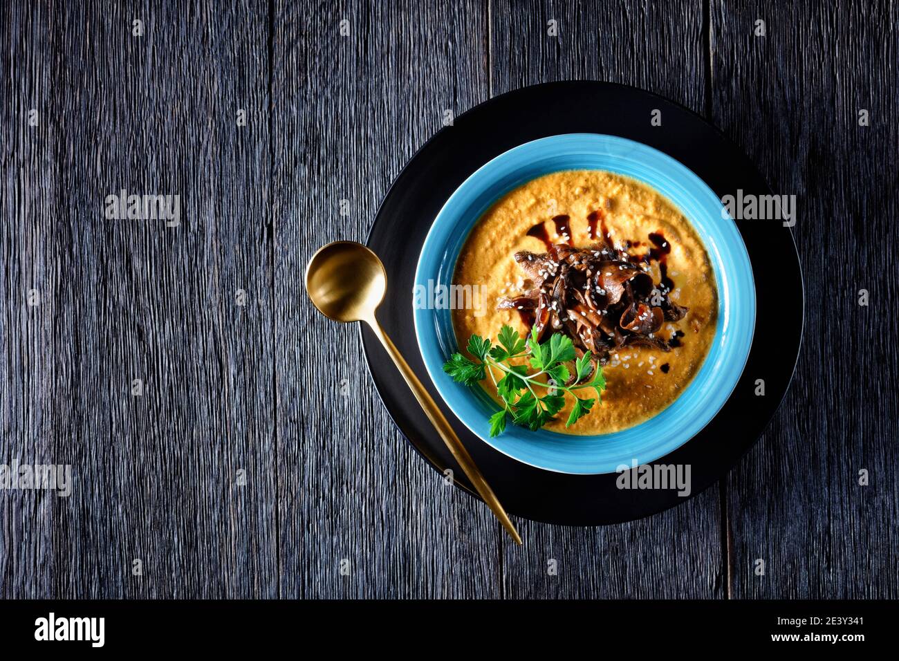 Zuppa di zucca di purea di zucca arrostita e brodo di funghi selvatici, con semi di sesamo, piatto vegano comfort, servita su un piatto blu con un cucchiaio su a. Foto Stock