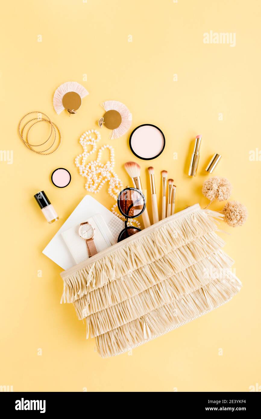 Borsa di paglia donna con accessori da viaggio, spazzole per il trucco, cosmetici, accessori dorati su sfondo giallo. Foto Stock