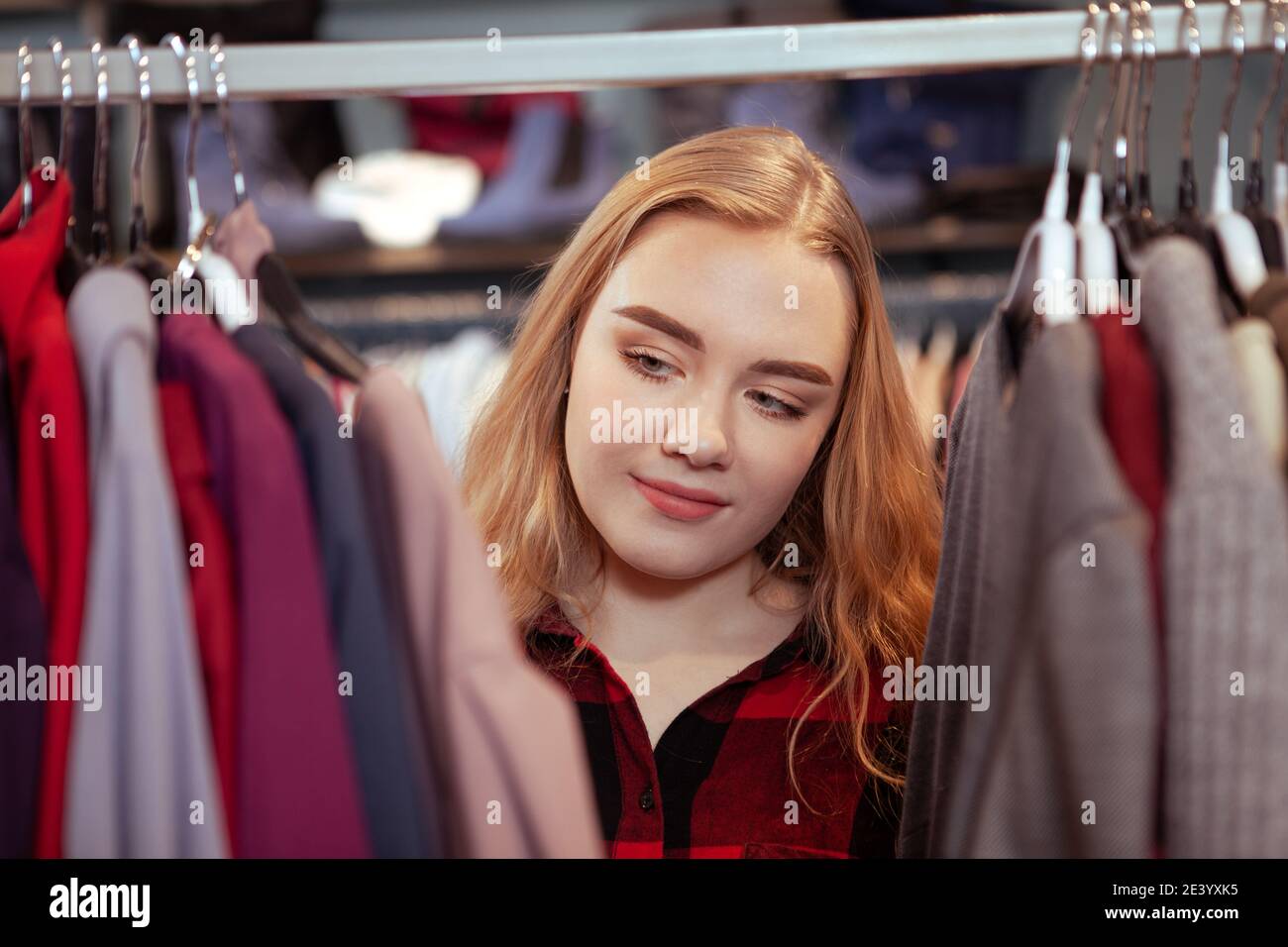 Giovane donna con capelli biondi che sceglie i vestiti da provare, facendo shopping al negozio di moda Foto Stock