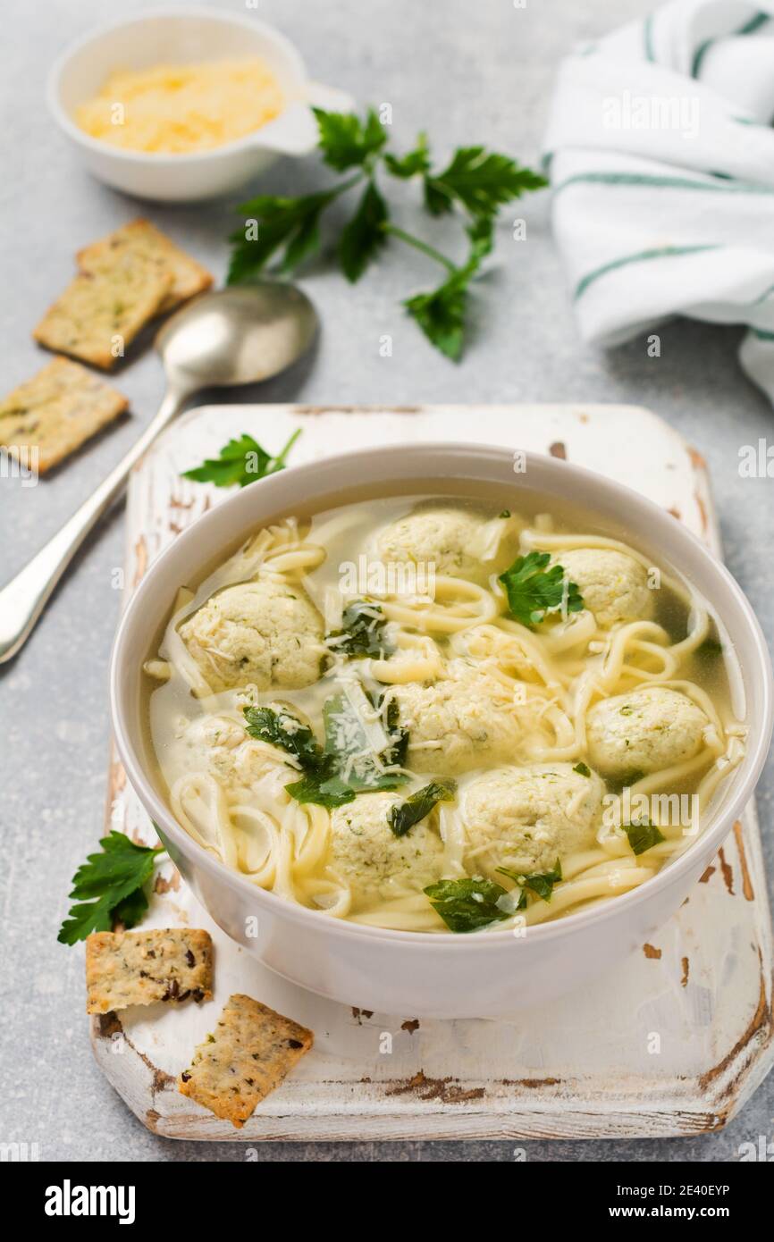 Ciotola di zuppa italiana tradizionale con polpette di pollo e pasta d'uovo, parmigiano, prezzemolo su sfondo grigio. Cucina italiana tradizionale. Foto Stock