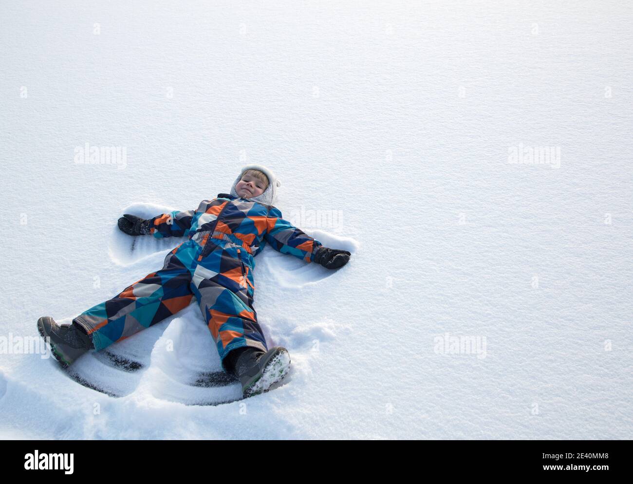 Ragazzo gioioso in tute invernali sdraiato sulla neve, muovendo la mano su e giù, giocando all'aperto, godendosi l'inverno. Un bambino nella neve mostra la fig Foto Stock